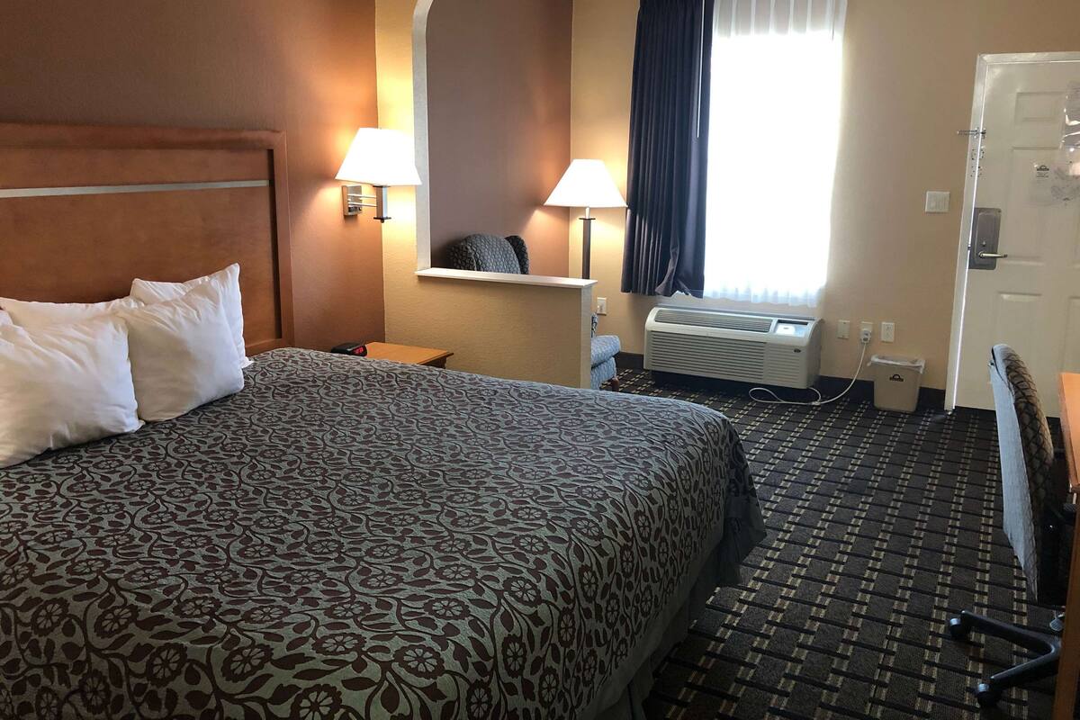 A hotel room with a large bed, two bedside lamps, a small table, a chair, a window with curtains, and an air conditioning unit.
