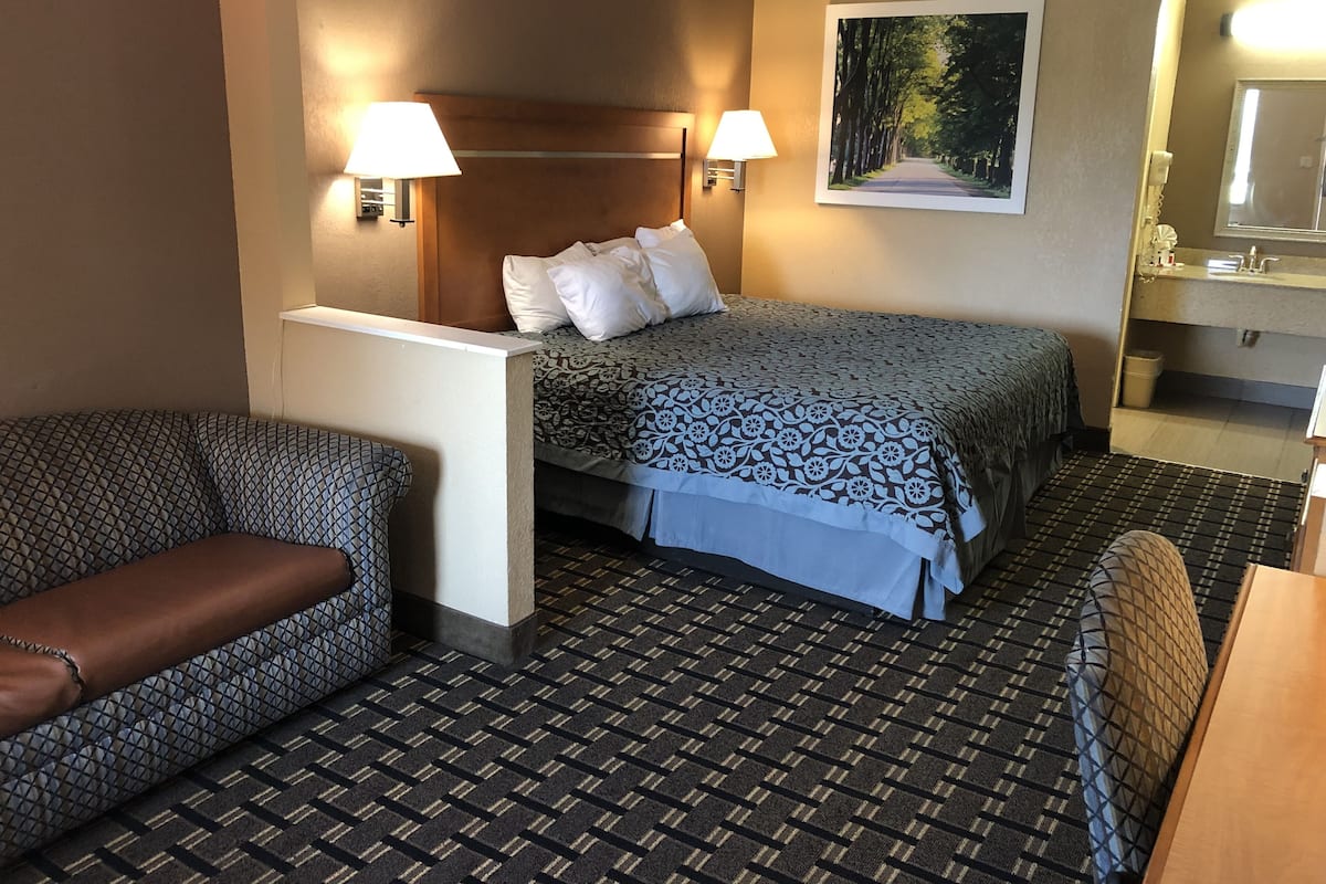 A hotel room with a bed, two bedside lamps, a painting on the wall, a bathroom with a sink and mirror, and patterned carpet.