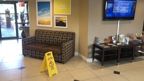 Ein Hotel-Rezeptionsbereich mit einer Couch, einem Fernseher der Wettervorhersage zeigt, gerahmten Bildern und einem Warnhinweis auf dem Boden.