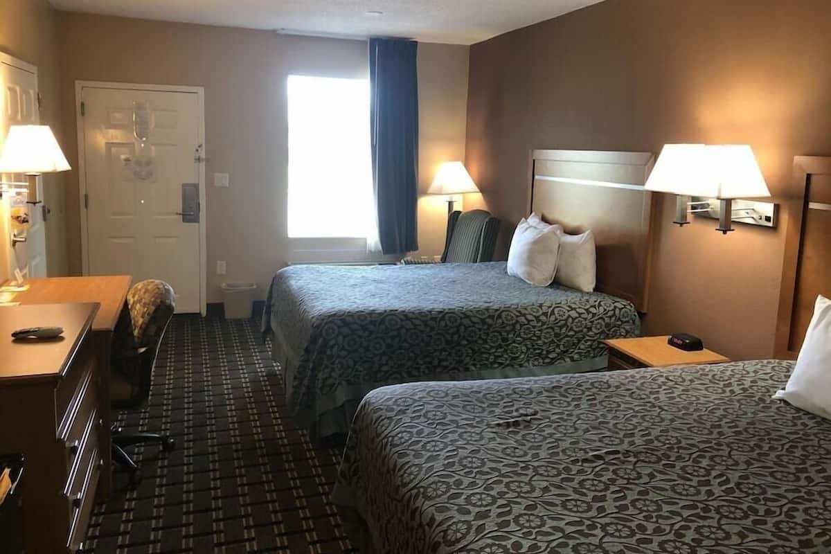 A hotel room with two beds, a desk, a chair, a window with curtains, and two wall lamps.