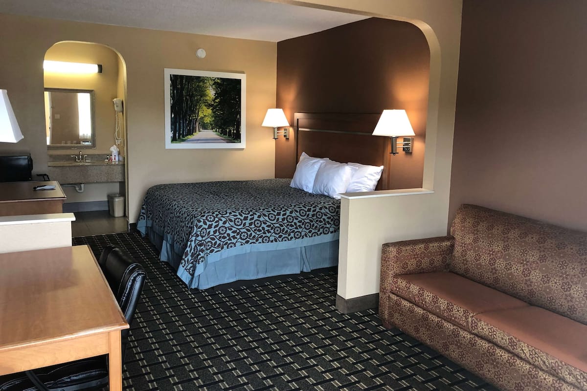 A hotel room with a bed, a desk, a chair, a sofa, a bathroom with a mirror, and a painting on the wall.