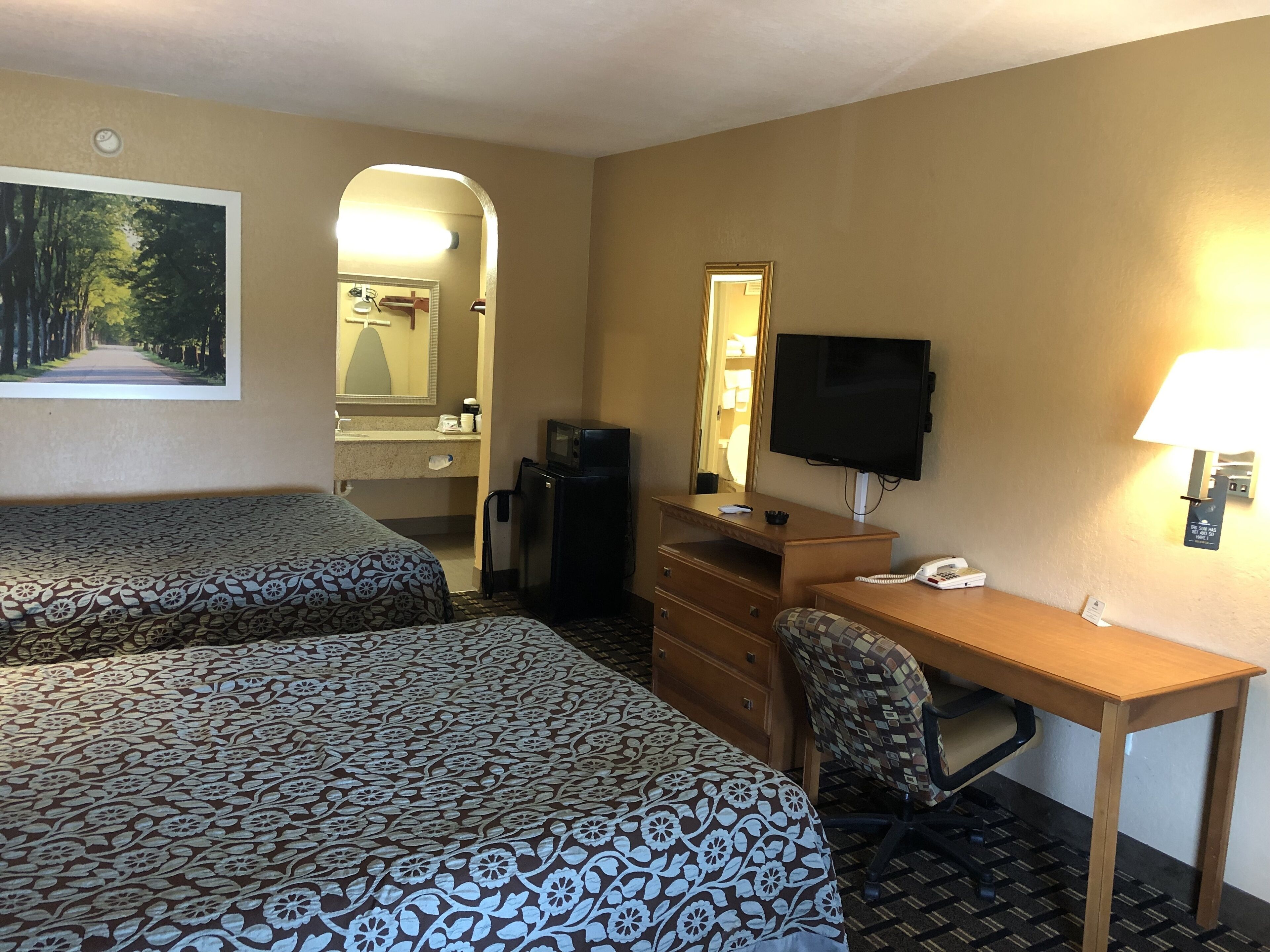 Une chambre d’hôtel avec deux lits, un bureau, une chaise, une télévision, un miroir, un réfrigérateur et une salle de bain visible à travers une arche.
