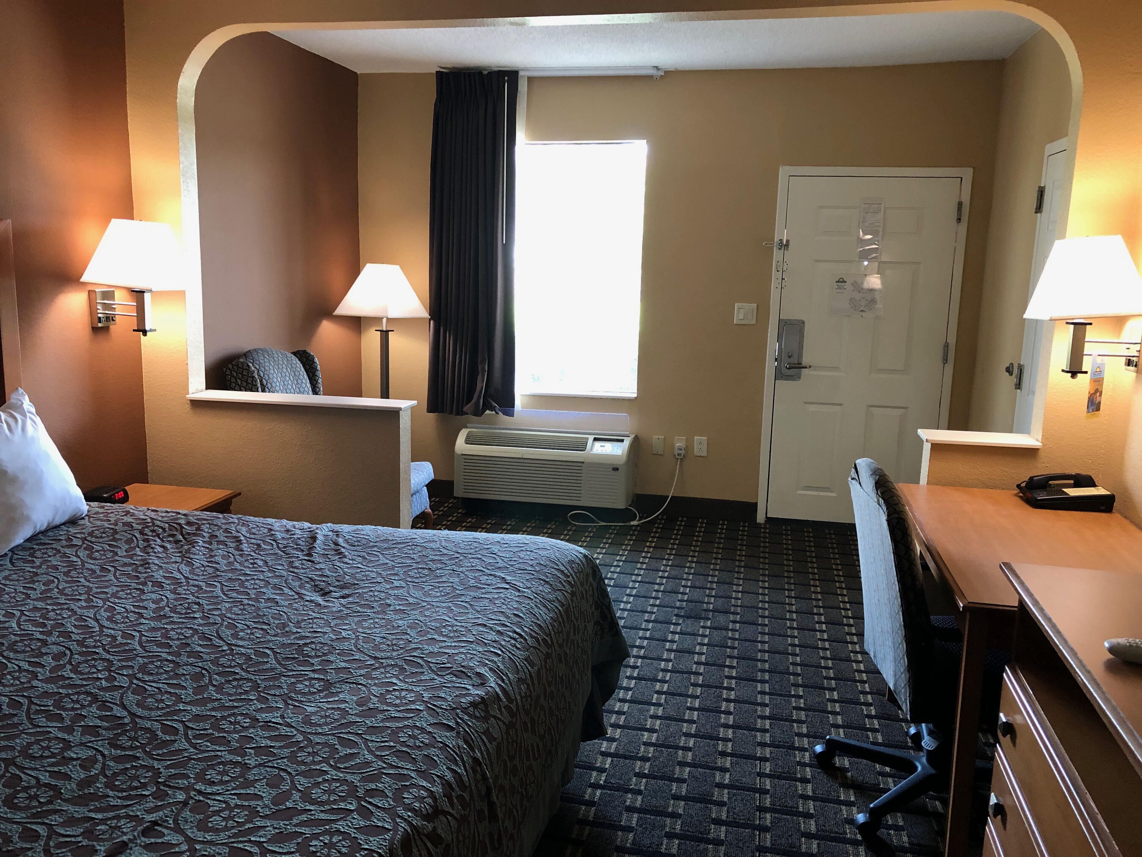 A hotel room with a large bed, two bedside lamps, a window with curtains, an air conditioning unit, a desk with a chair, and a door with a lock.