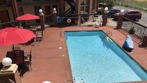 Ein rechteckiger Pool mit Sprungverbotsschild, umgeben von einer roten Terassenfliese, Sitzgelegenheiten im Freien und einem überdachten Bereich mit Sonnenschirmen.