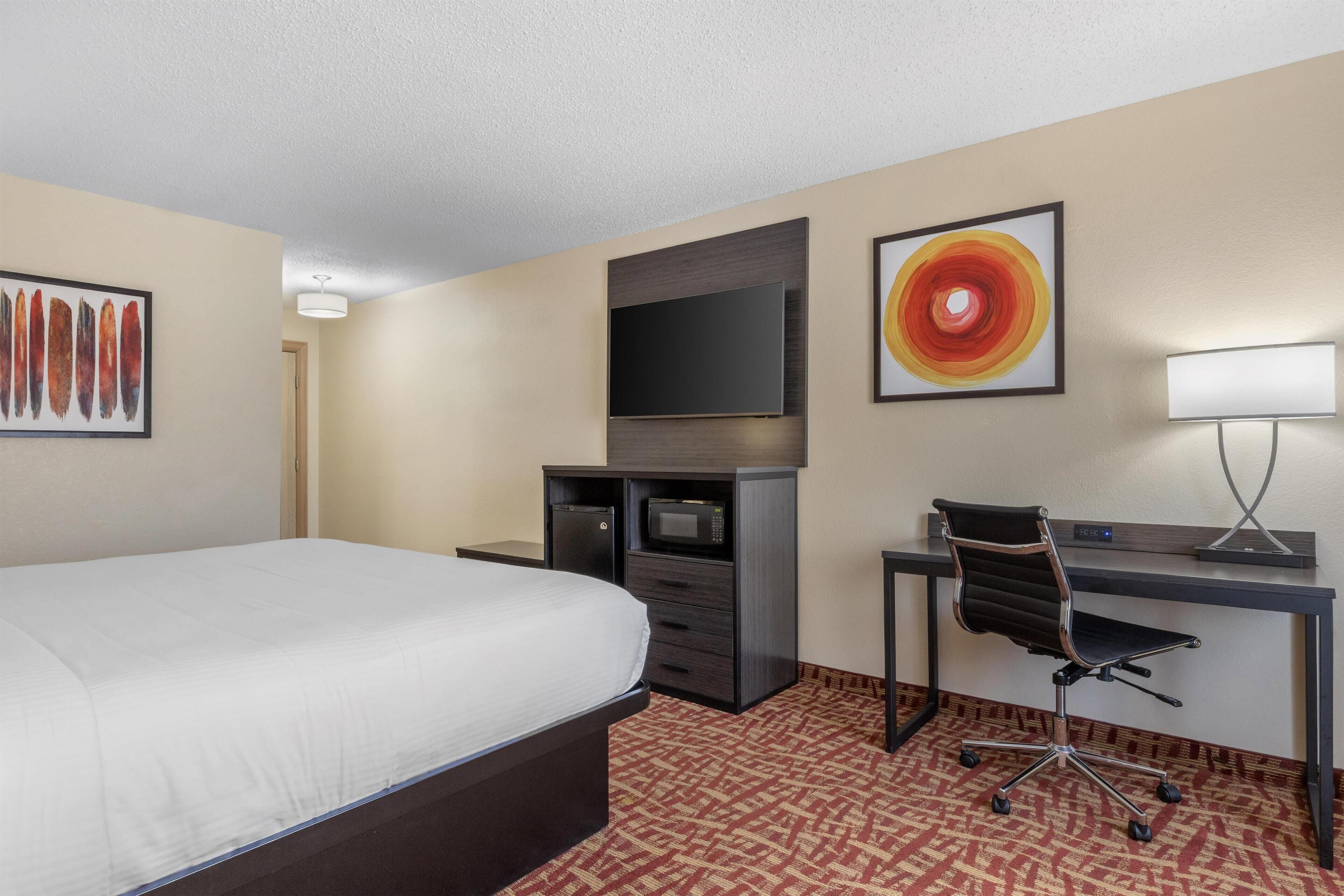 A hotel room with a bed, a desk with a chair, a television, a microwave, and artwork on the walls.