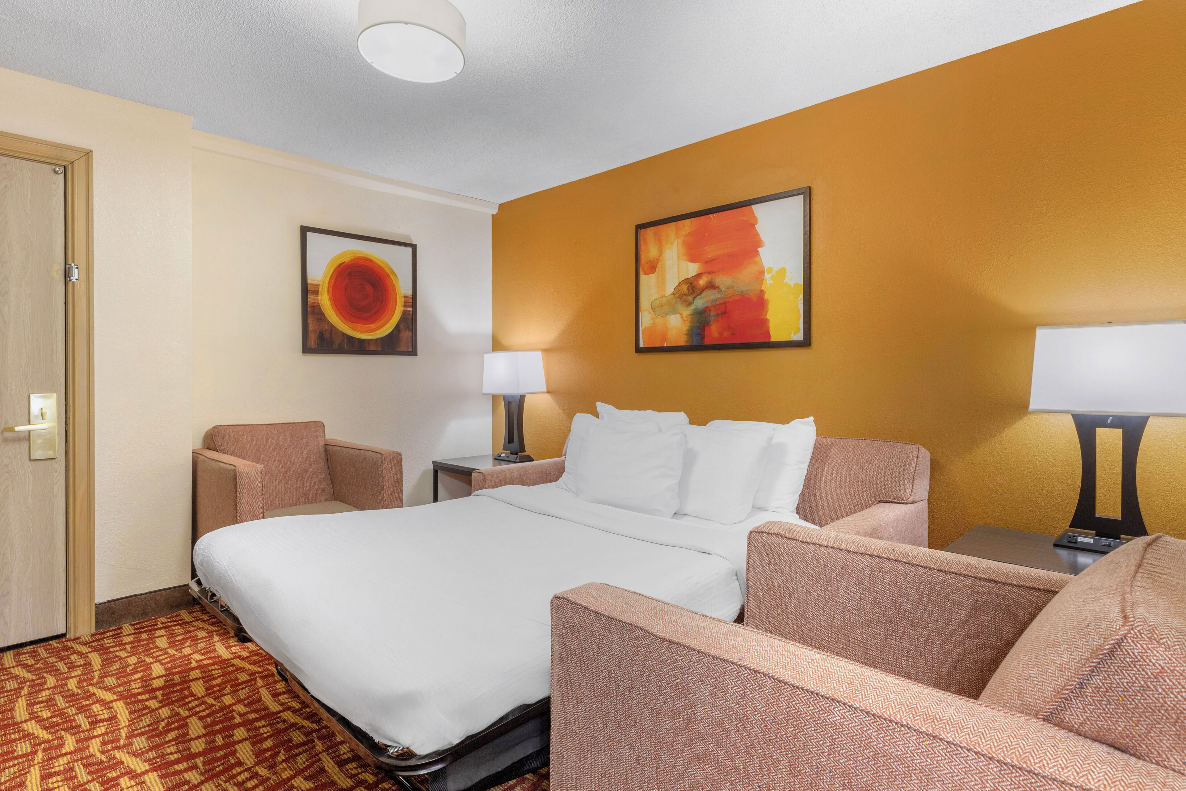 Habitación de hotel con una cama, dos sillones, una mesita de noche, una lámpara y dos cuadros en la pared.