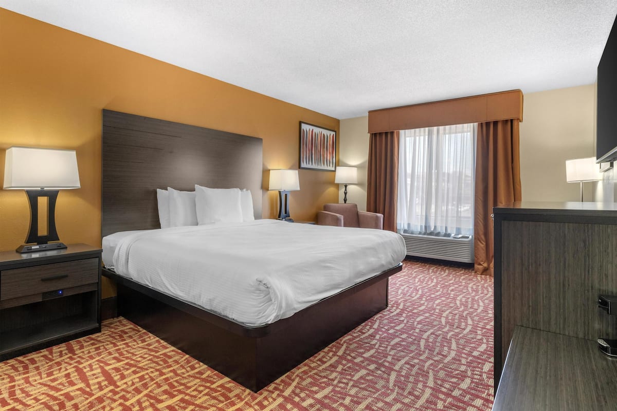 A hotel room with a large bed, two bedside tables with lamps, a desk, a chair, a TV, and a window with curtains.
