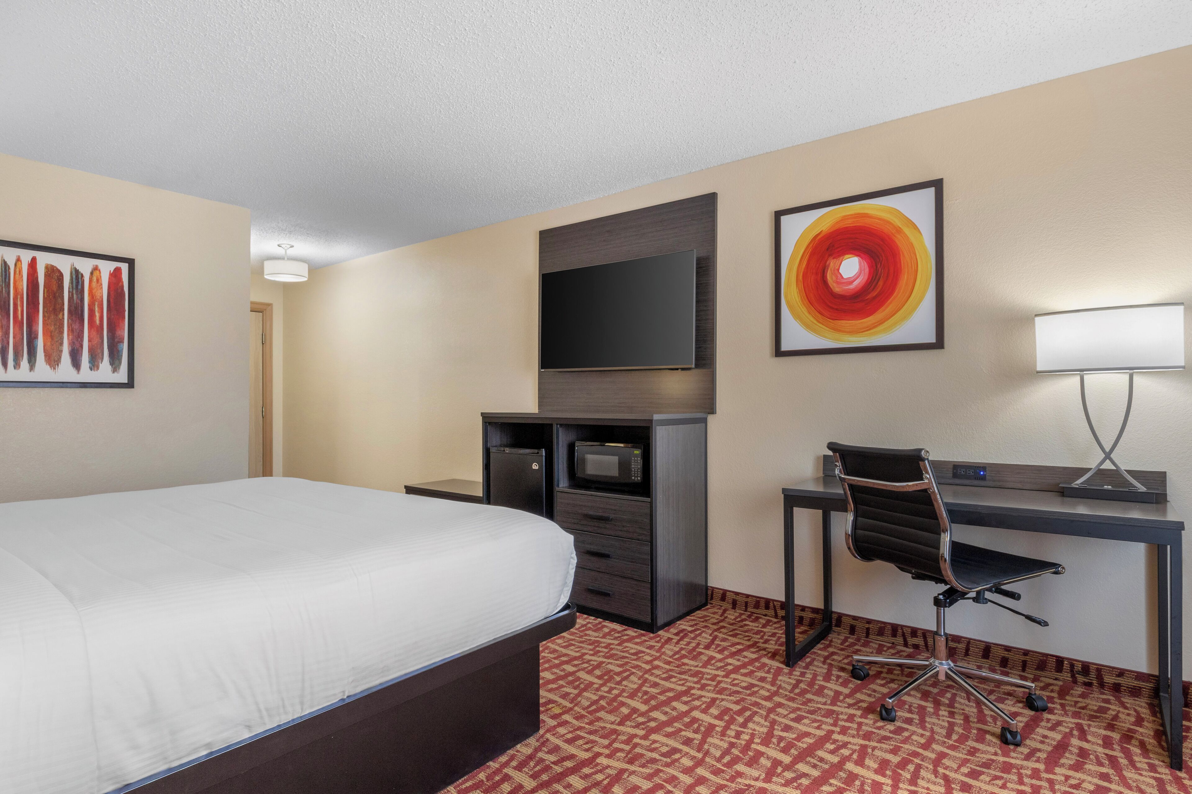 A hotel room with a bed, a desk with a chair, a flat-screen TV, a microwave, and artwork on the walls.