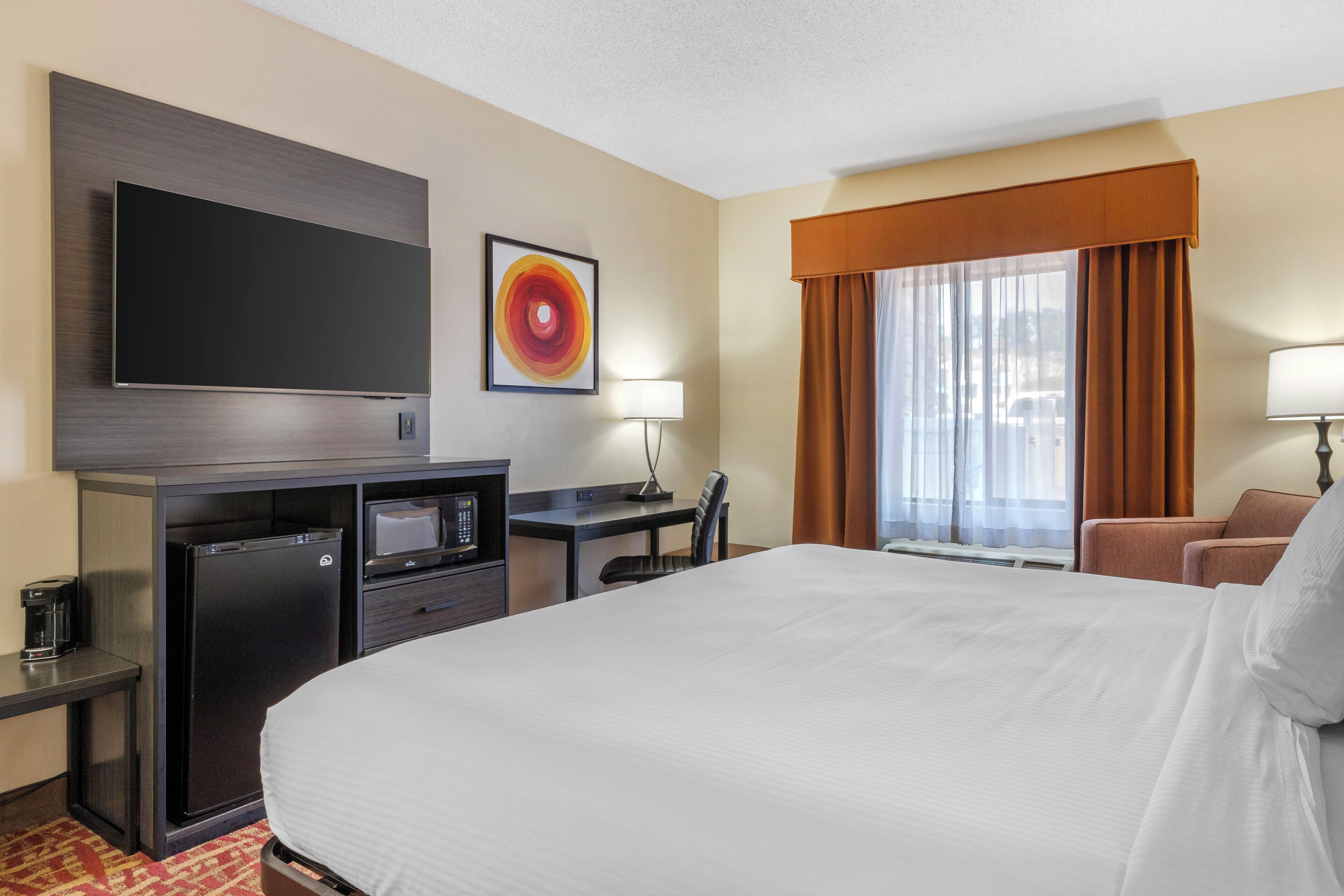 A hotel room with a large bed, a desk with a computer, a flat-screen TV, a refrigerator, and a window with curtains.
