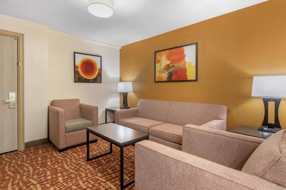 A hotel room with a beige sofa, a chair, a small table, and two lamps. There are two paintings on the walls.