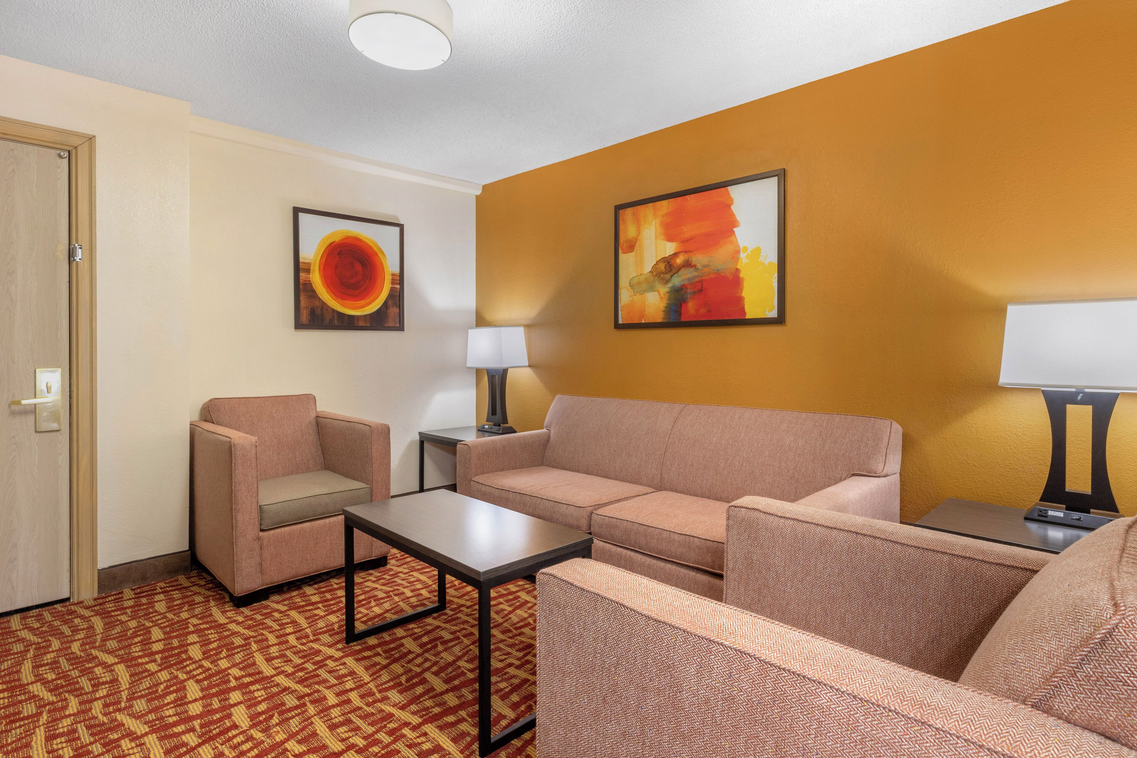 A hotel room with a beige sofa, a matching armchair, a small table, and two lamps. There are two framed artworks on the walls.