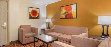 A hotel room with a beige sofa, a matching armchair, a small table, and two lamps. There are two framed artworks on the walls.
