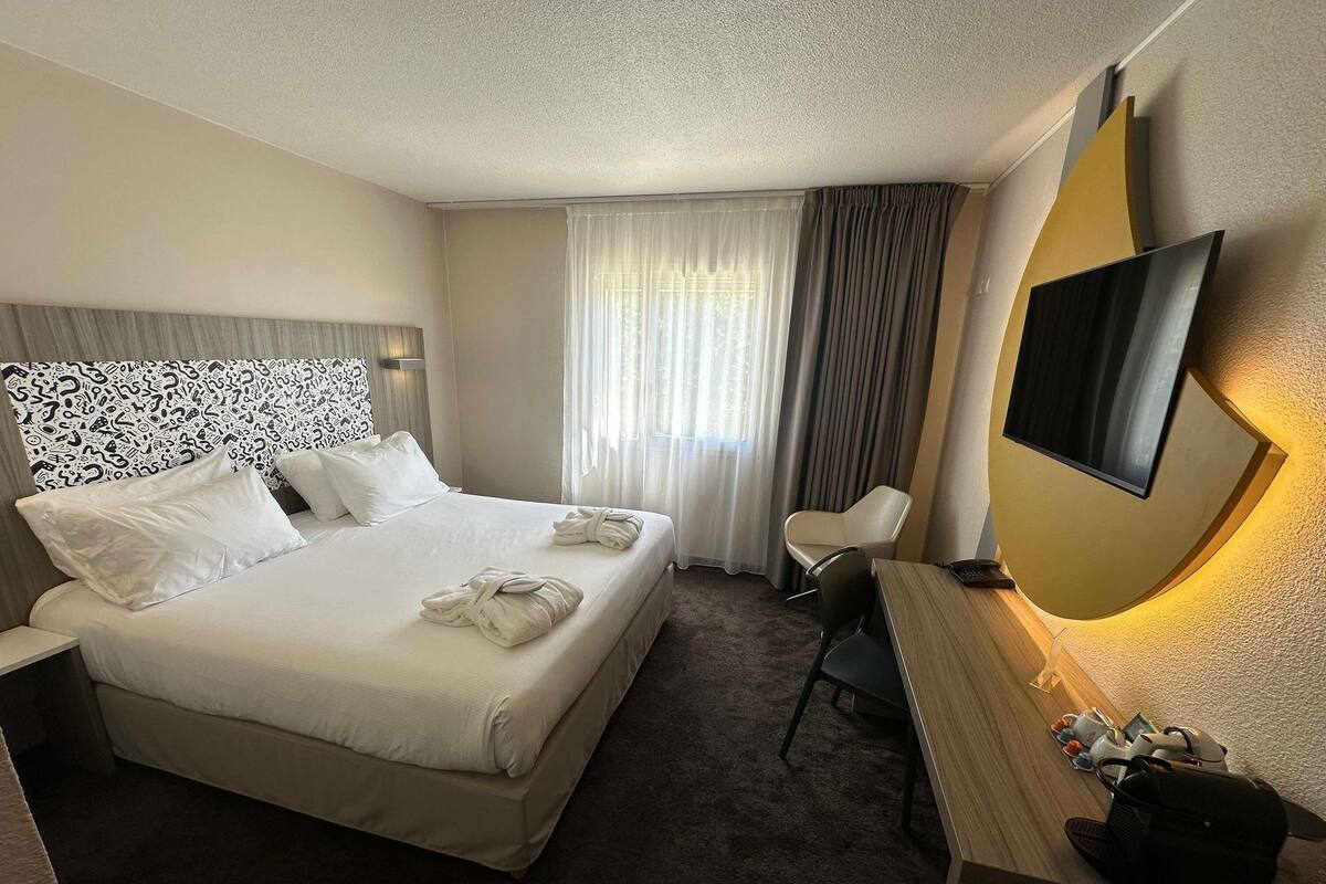 Habitación de hotel con una cama grande, un escritorio, una silla, un televisor y una ventana con cortinas.