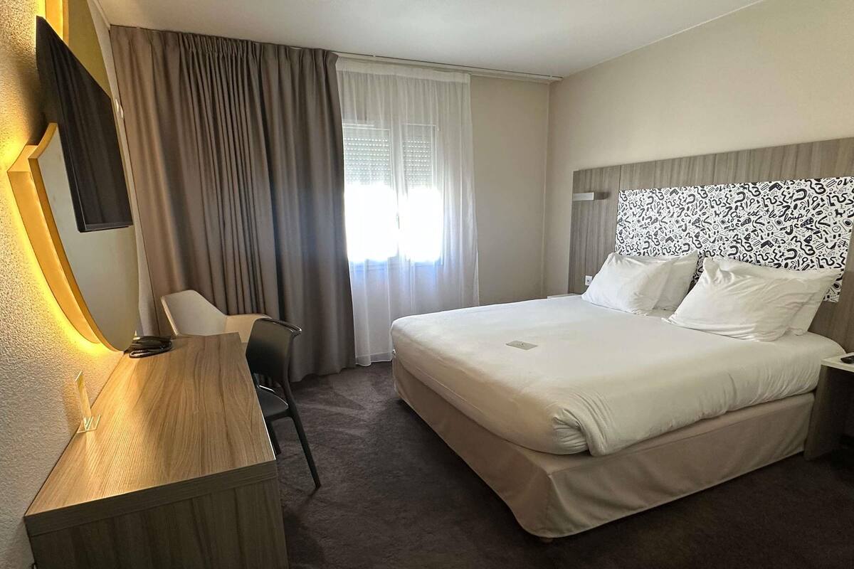 Una habitación de hotel con cama, escritorio, silla y ventana con cortinas.