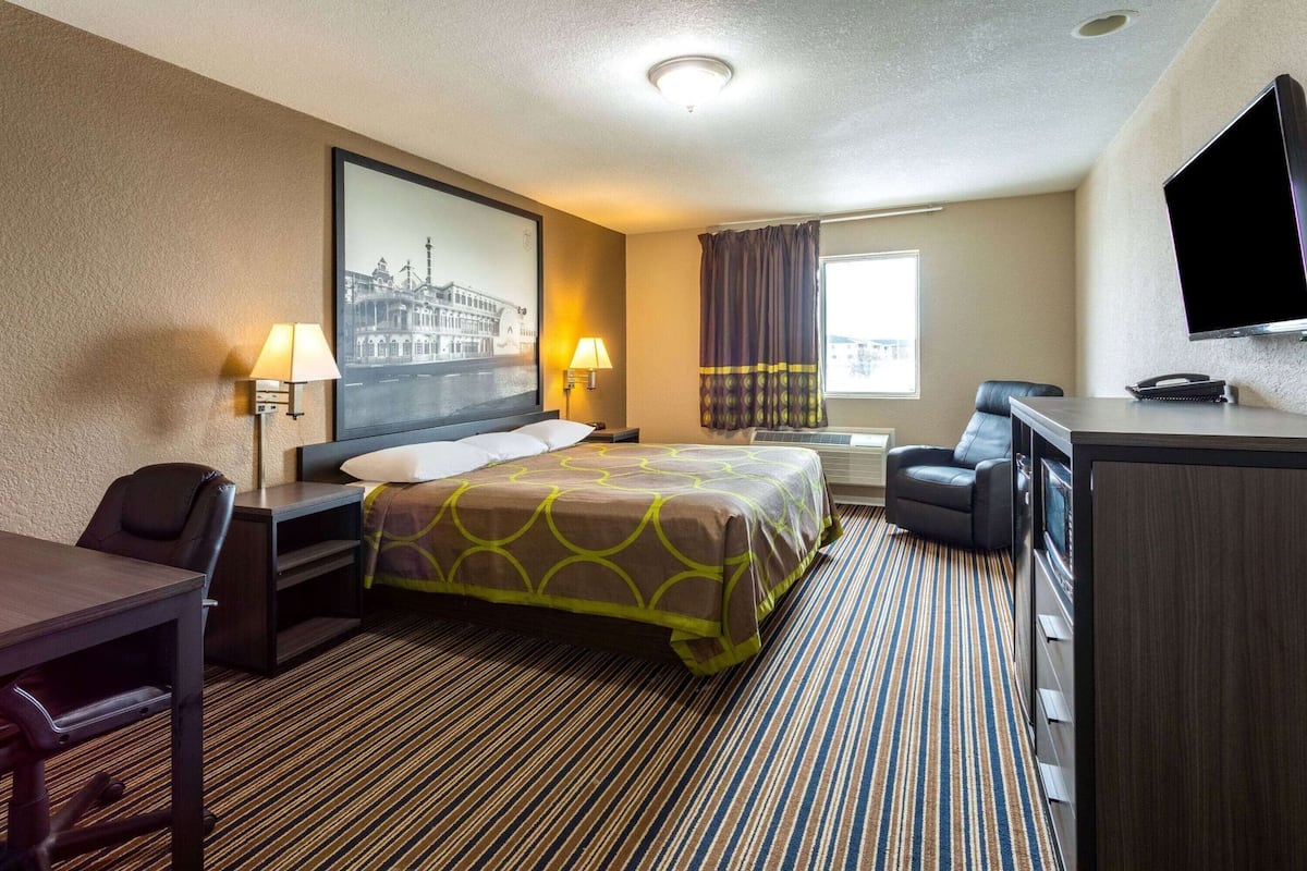 A hotel room with a bed, desk, chair, TV, and a large window with curtains.