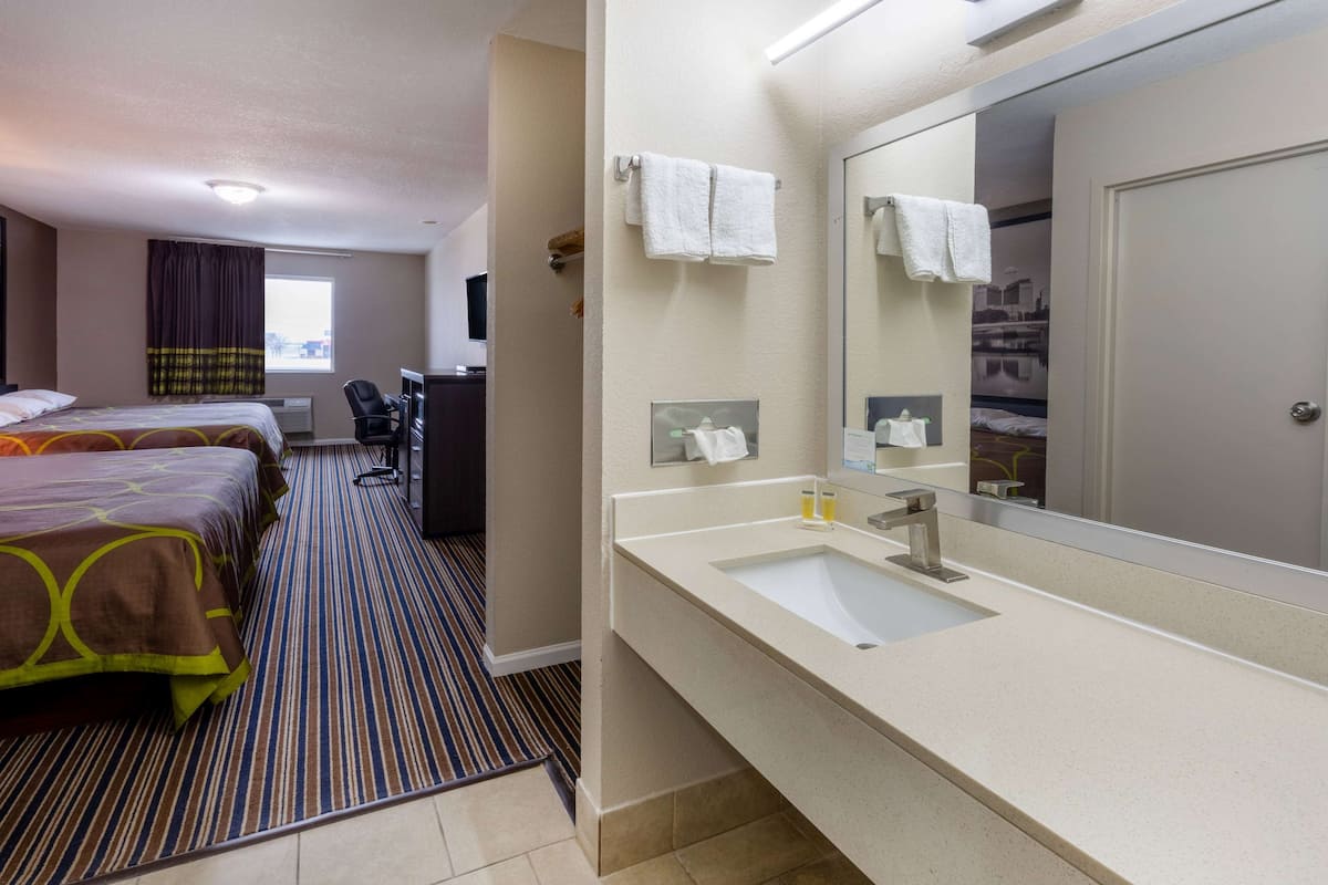 A hotel room with two beds, a desk, a large mirror, and a bathroom with a sink and towel rack.