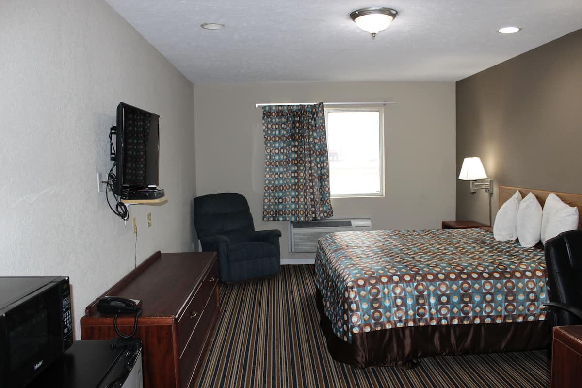 A hotel room with a bed, a desk with a telephone, a chair, a television mounted on the wall, and a window with curtains.