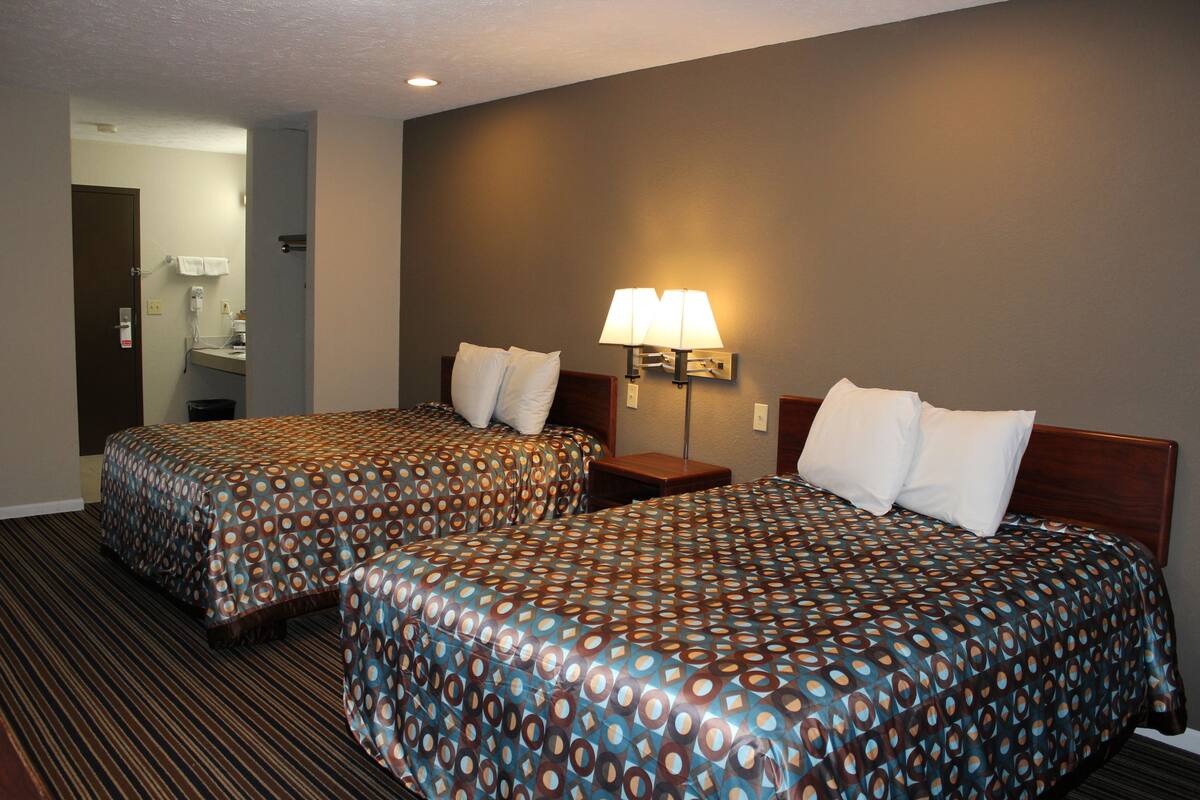 A hotel room with two beds, a nightstand, a lamp, and a bathroom visible through an open door.