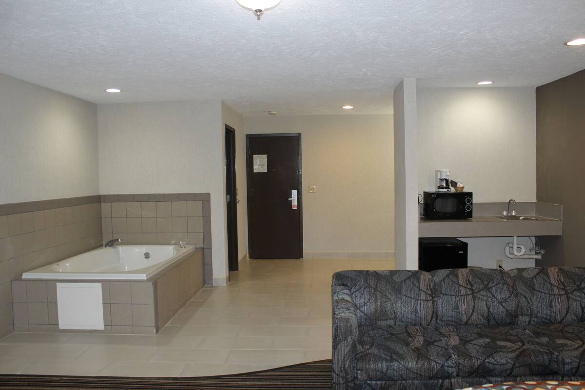 A hotel room with a large bathtub, a sofa, a kitchenette with a microwave, and a door leading to another room.