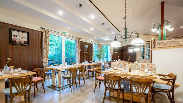 Un restaurante con mesas y sillas de madera, ventanas amplias con cortinas, y lámparas colgantes.
