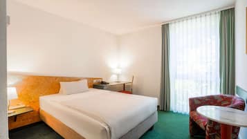 A hotel room with a large bed, a desk with a lamp, a chair, and a window with curtains.