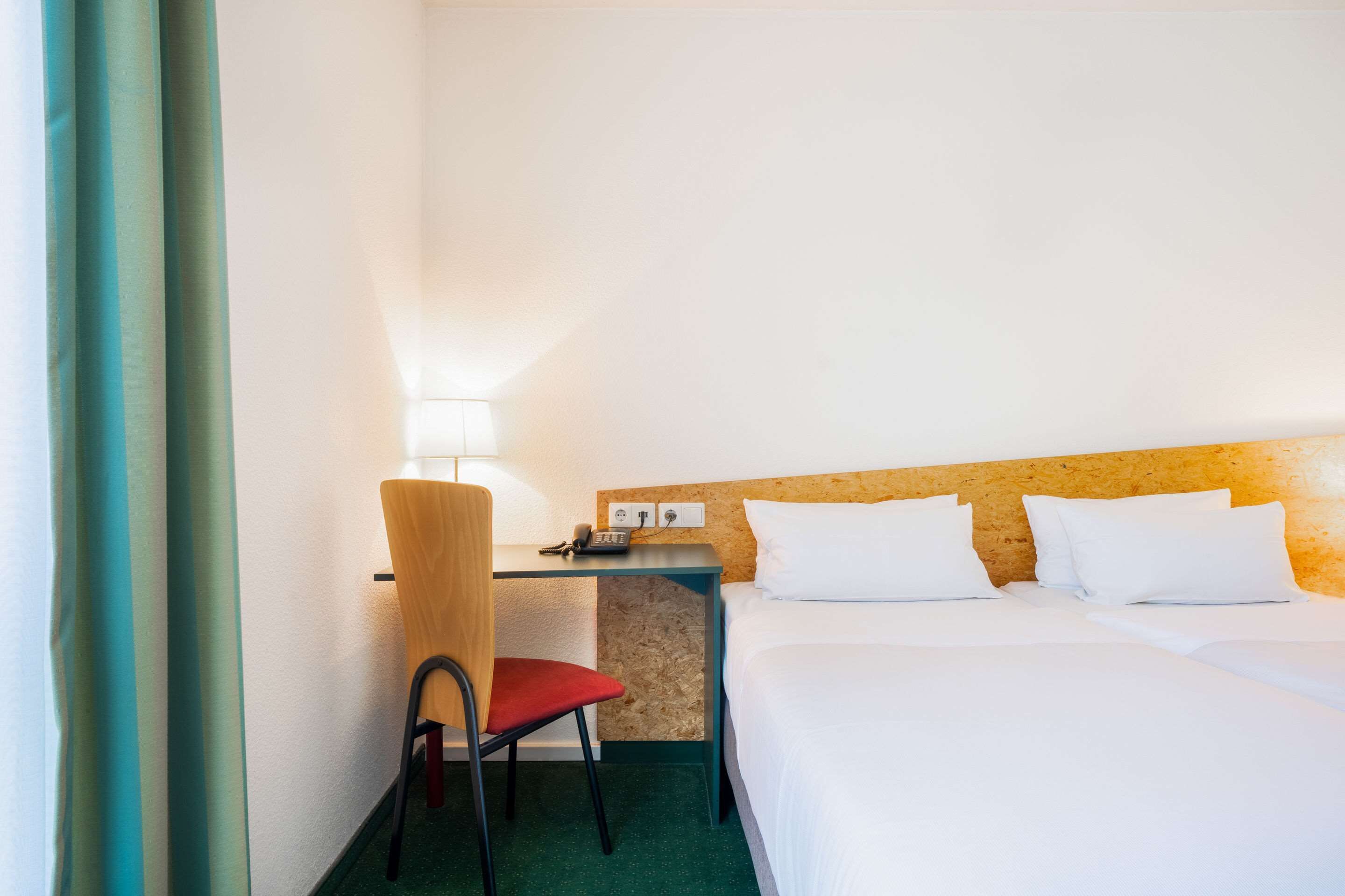 Una habitación de hotel con una cama, un escritorio con silla, una lámpara y un enchufe.