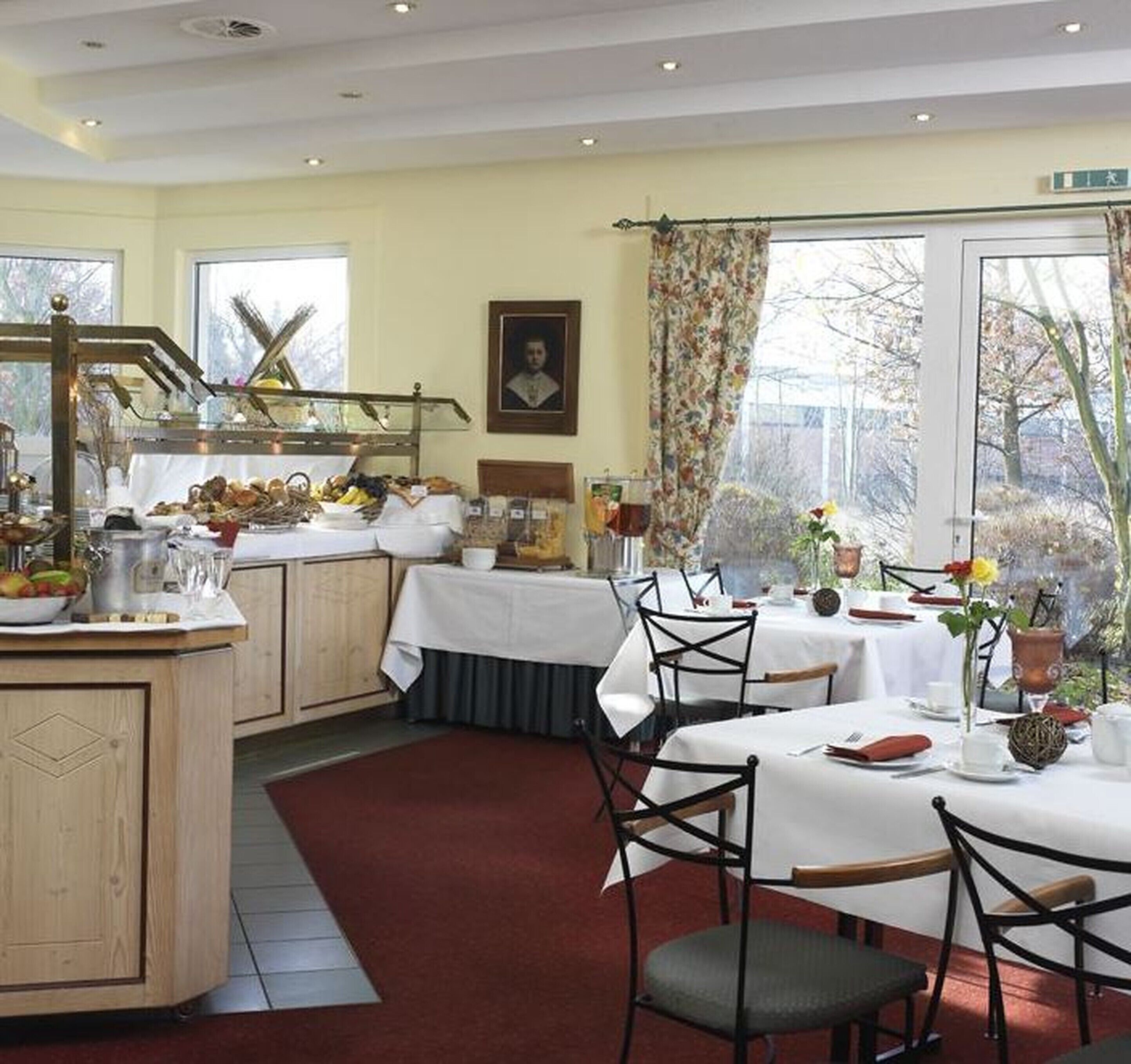 A dining area with a central island, white tablecloths, and chairs. There is a large window with a view of trees and a door with a sign above it.