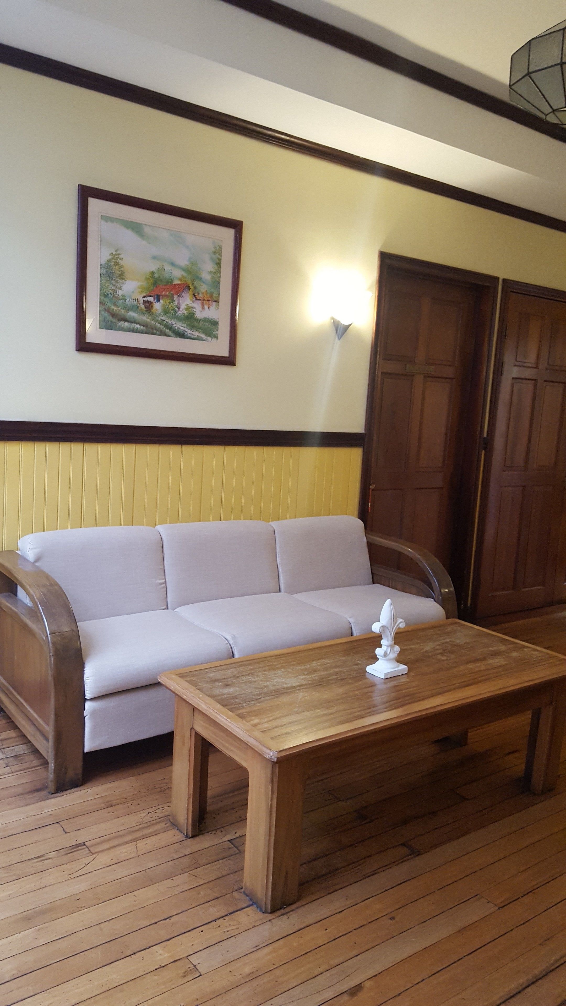 A room with a wooden floor, a beige sofa, a wooden coffee table, a framed painting, and a wall-mounted light fixture.