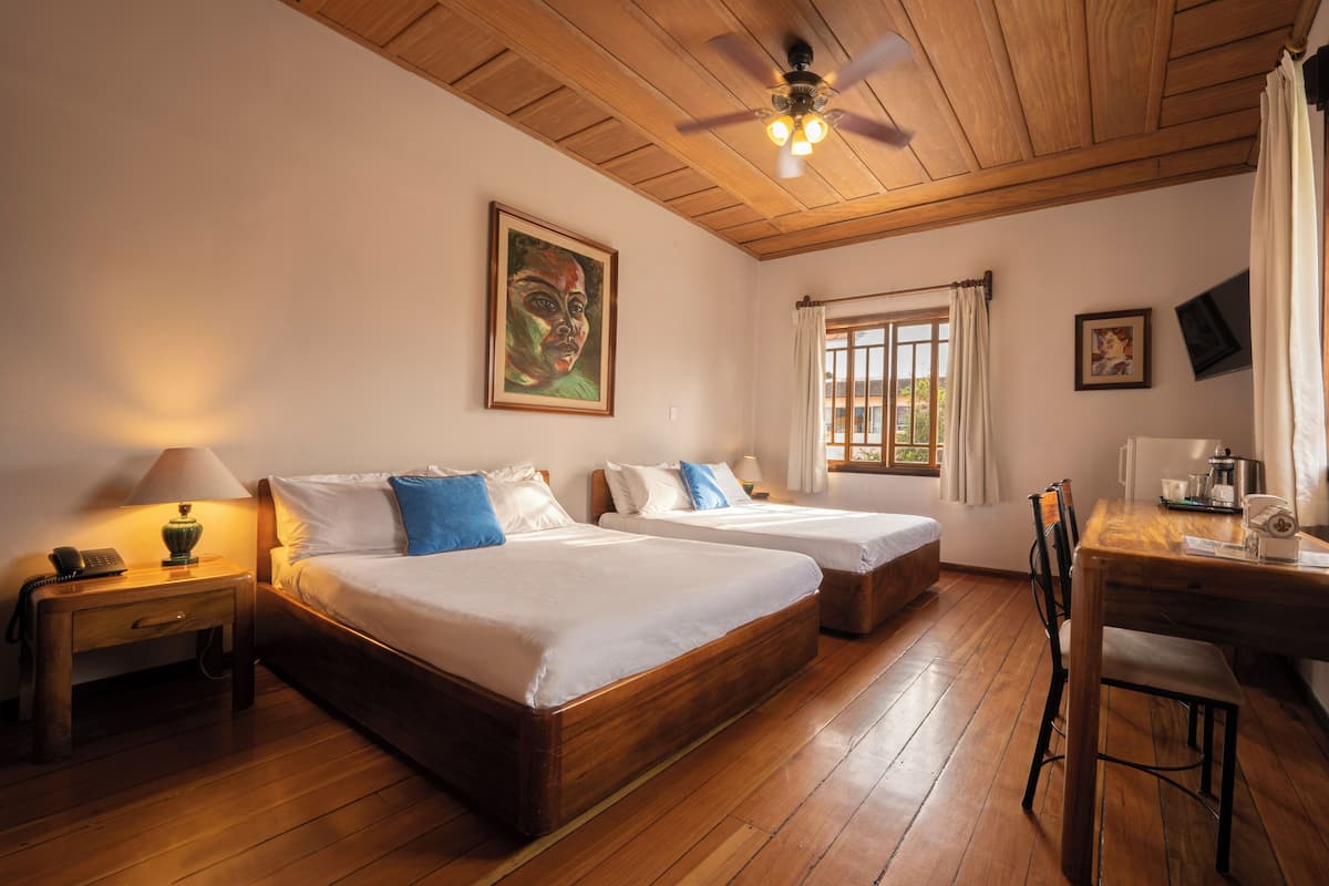 A room with two beds, a wooden ceiling, a window with curtains, a desk with a chair, and a painting on the wall.