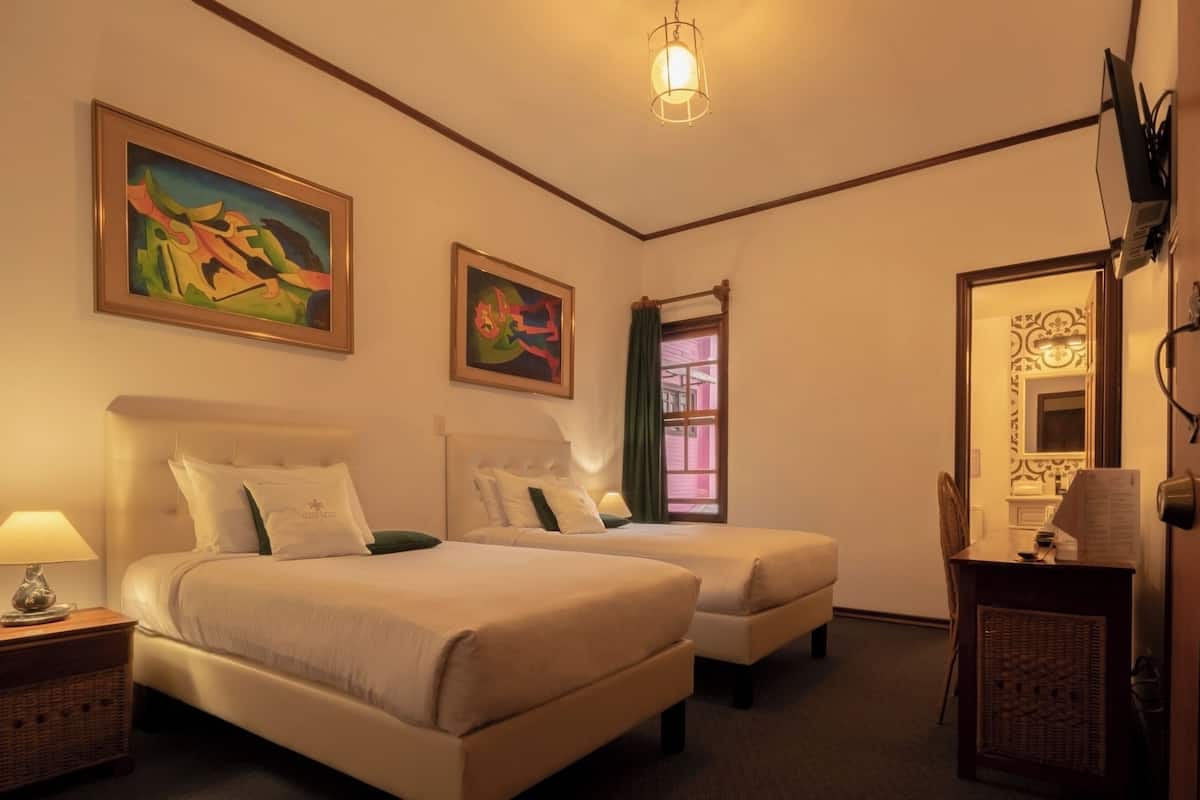 A hotel room with two beds, a desk, a lamp, and artwork on the walls.