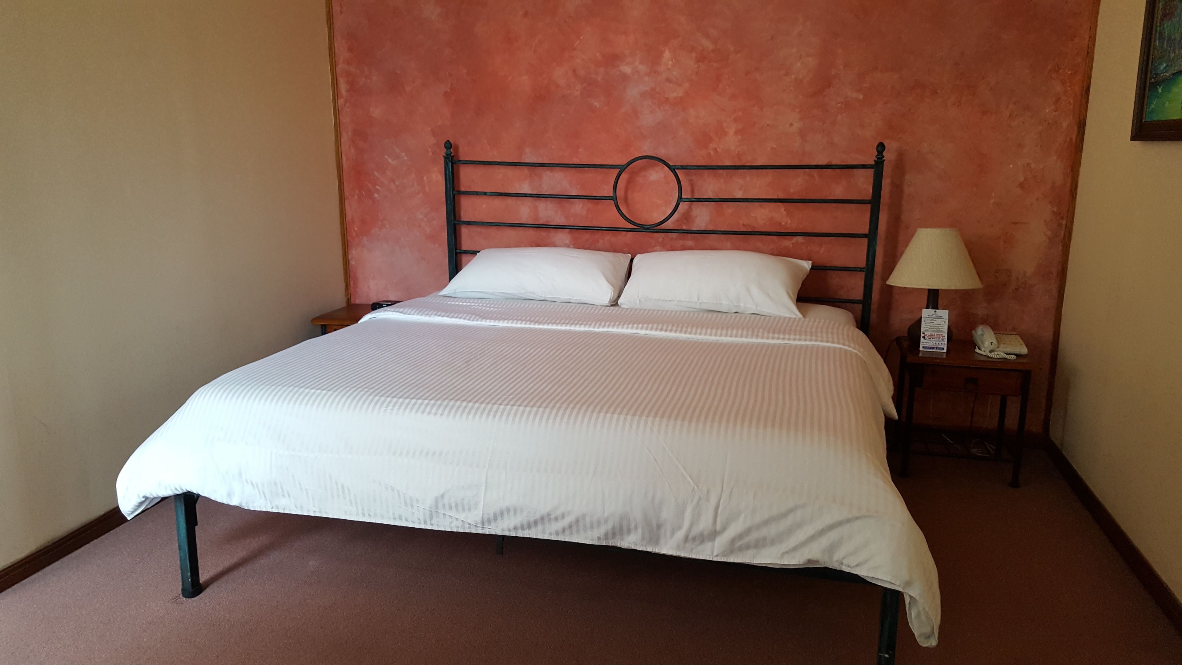 Una cama con cabecera metálica, ropa de cama blanca y una mesita de noche con una lámpara y un teléfono.