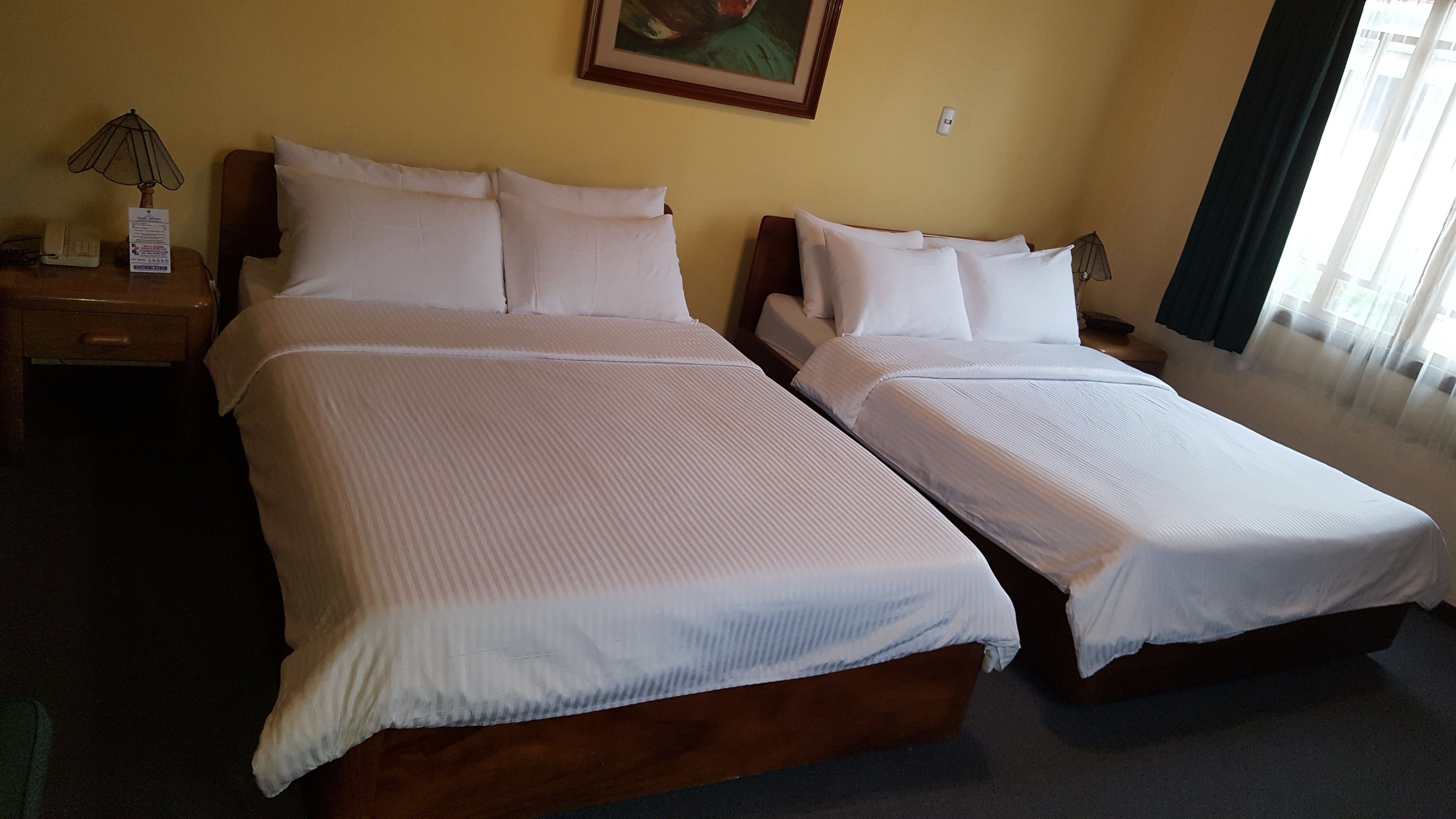 A hotel room with two single beds, a nightstand with a lamp, a painting on the wall, and a window with curtains.
