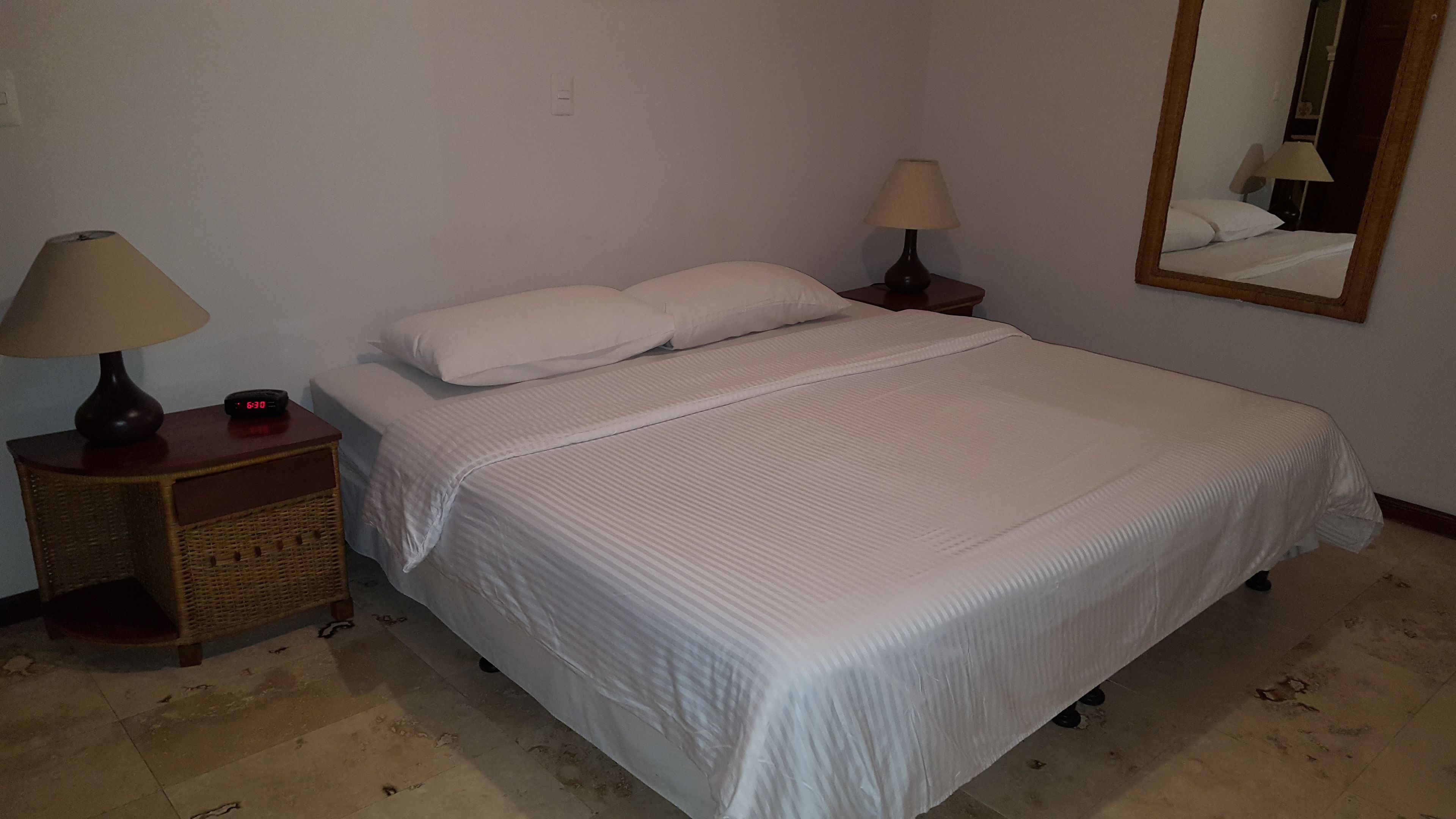 A neatly made bed with white bedding, two bedside lamps, a digital clock, and a wicker nightstand.