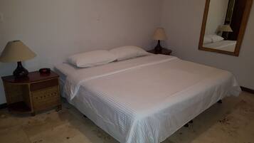 A neatly made bed with white bedding, two bedside lamps, a digital clock, and a wicker nightstand.