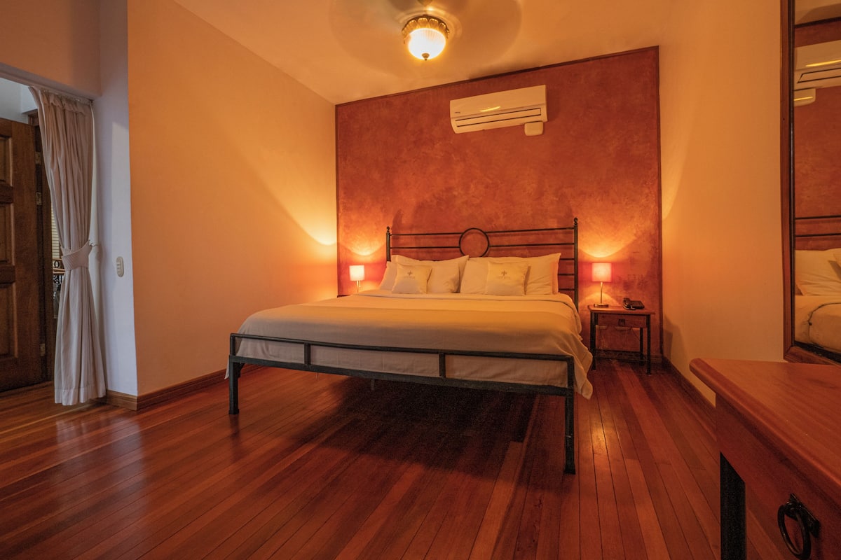 A bedroom with a wooden bed, an air conditioning unit, a nightstand with a lamp, and a wooden door.
