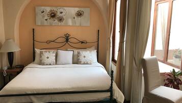 Una cama bien hecha con ropa blanca y almohadas decorativas, una lámpara de noche y un cuadro de flores en la pared.