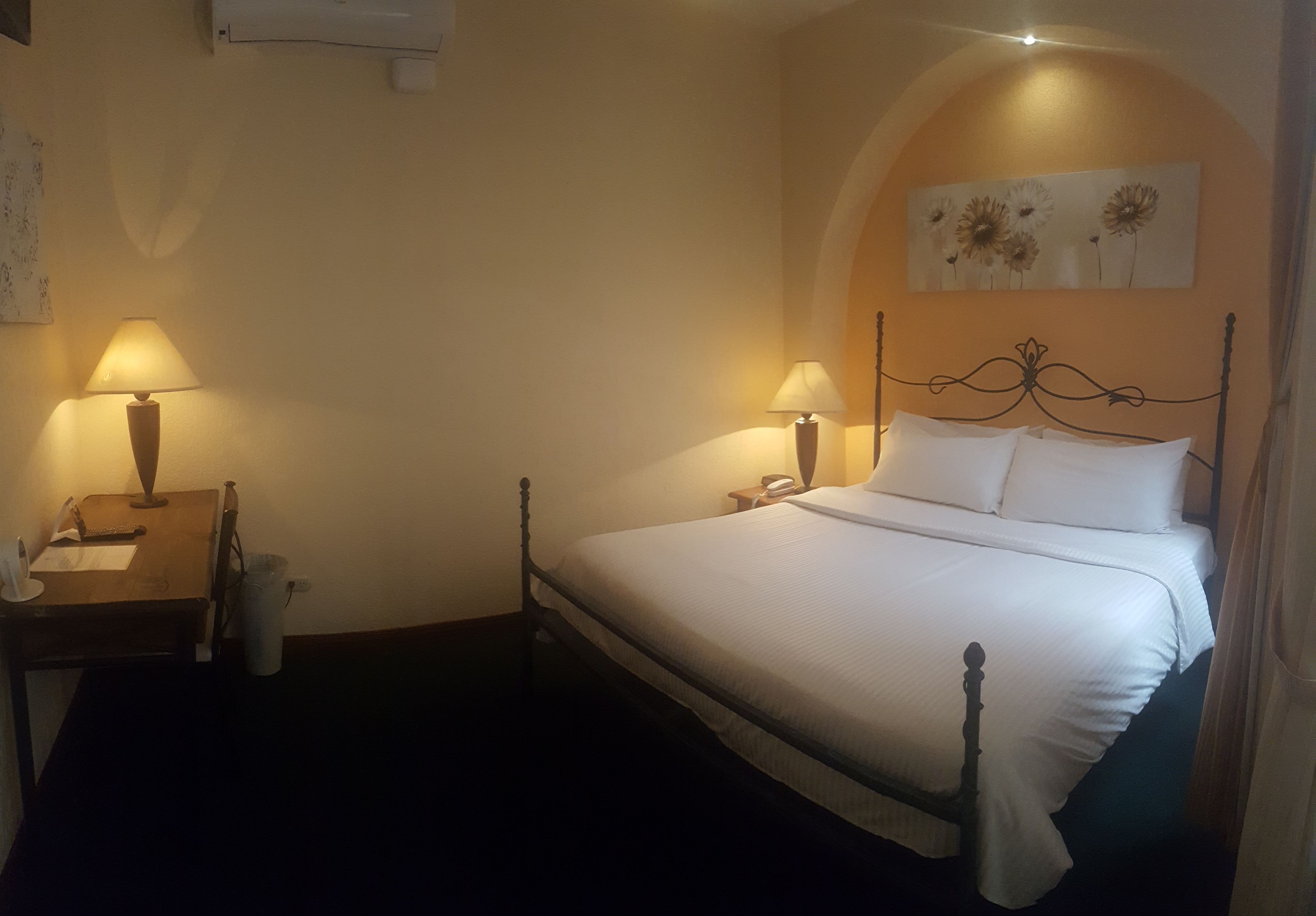 Uma cama bem arrumada com lençóis brancos, duas luminárias de cabeceira, uma mesinha com abajur e um ar-condicionado fixado na parede.