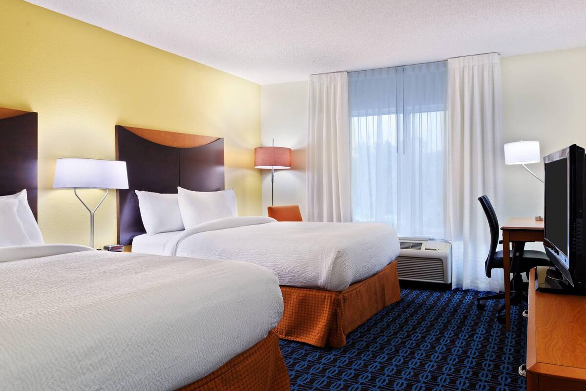 A hotel room with two beds, a desk with a computer, a chair, a lamp, and a window with curtains.