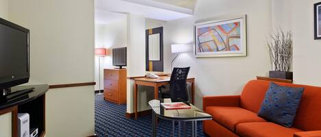 Een hotelkamer met een televisie, een bureau met een computer, een stoel, een oranje bank en een kleine tafel.