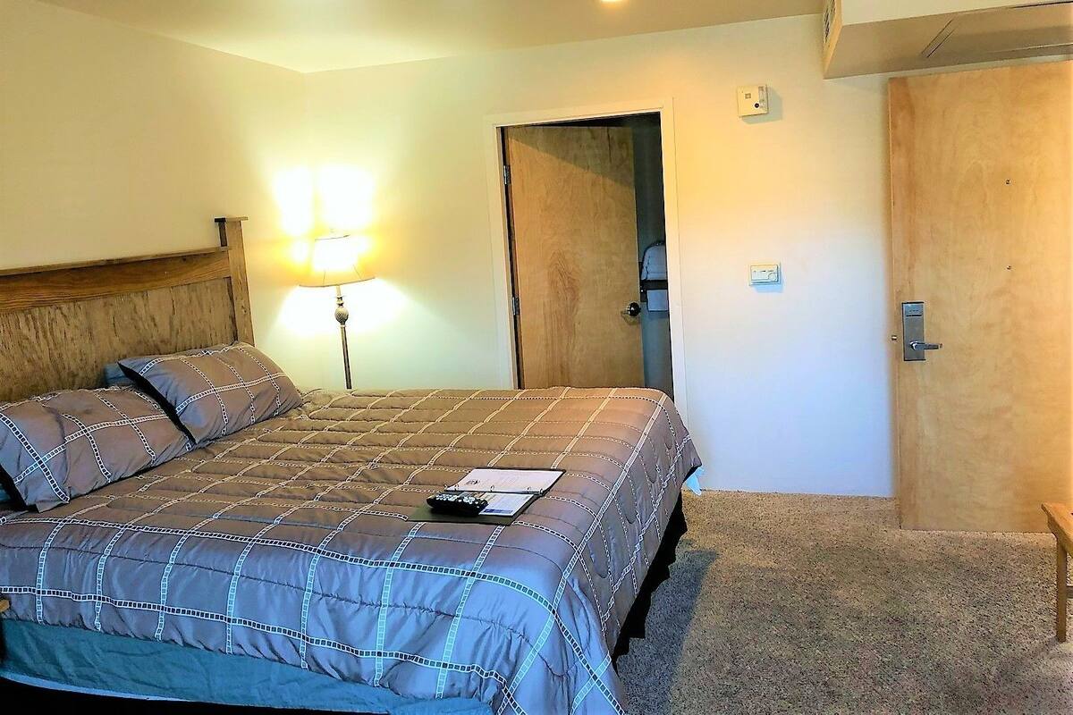 A hotel room with a large bed, a bedside lamp, a door, and a small table with a remote control.