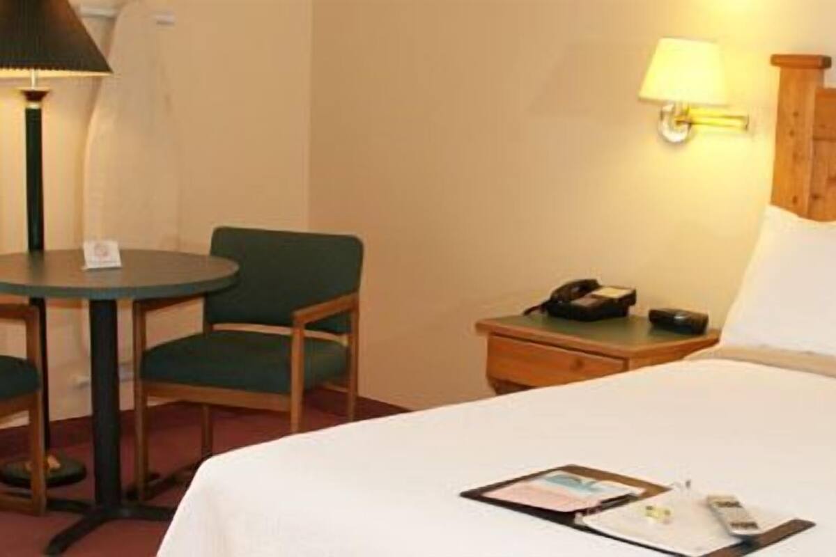 A hotel room with a bed, a desk with two chairs, a lamp, a nightstand, and a wall-mounted lamp.