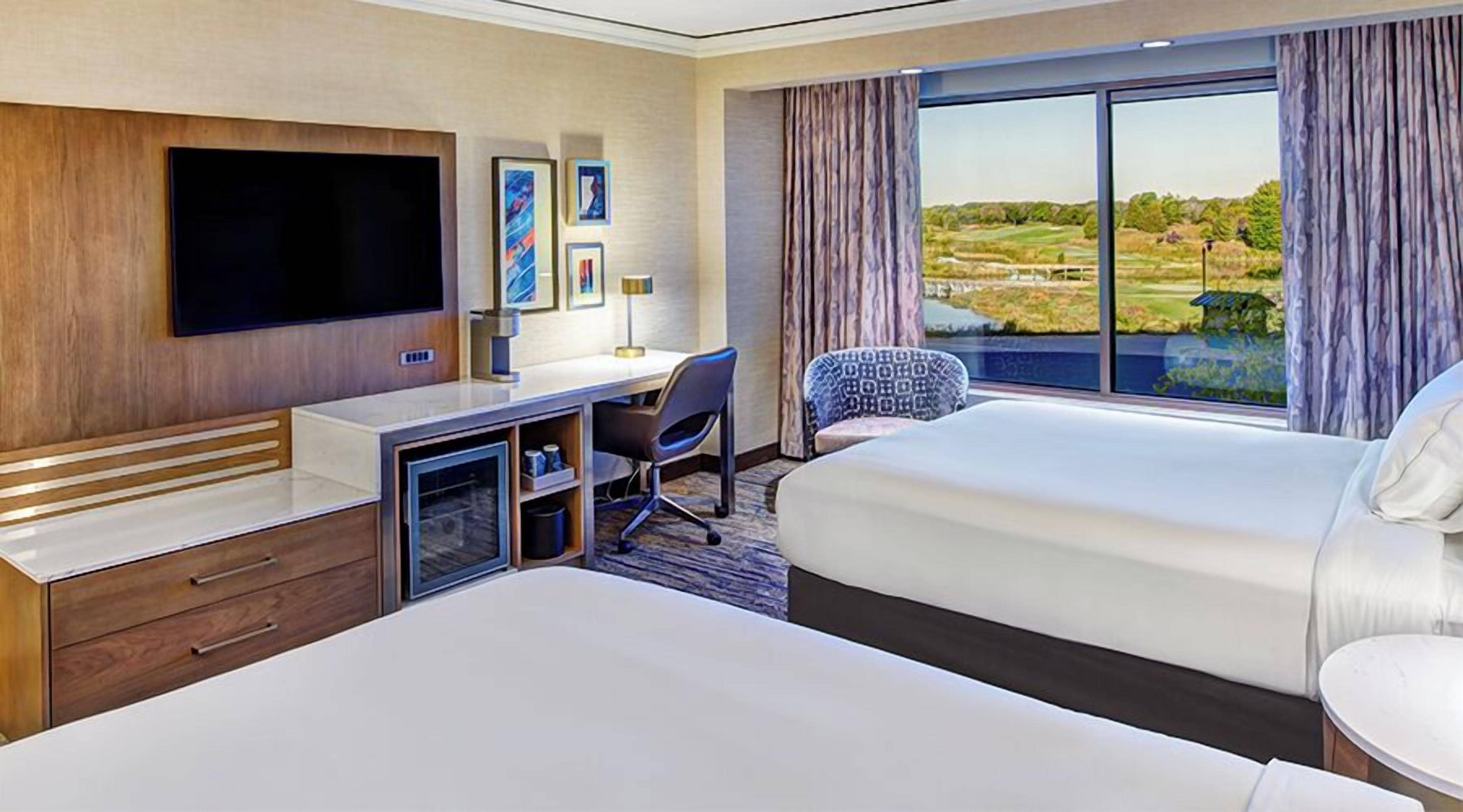 A hotel room with a large bed, a desk with a chair, a flat-screen TV, and a large window with a view of a golf course.