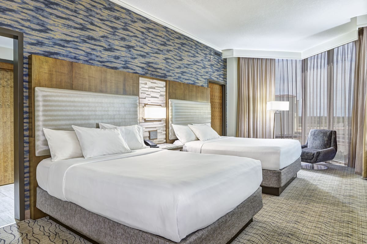 Ein Hotelzimmer mit zwei Betten, einem großen Fenster mit Vorhängen, einem Sessel und einem gemusterten Teppich.