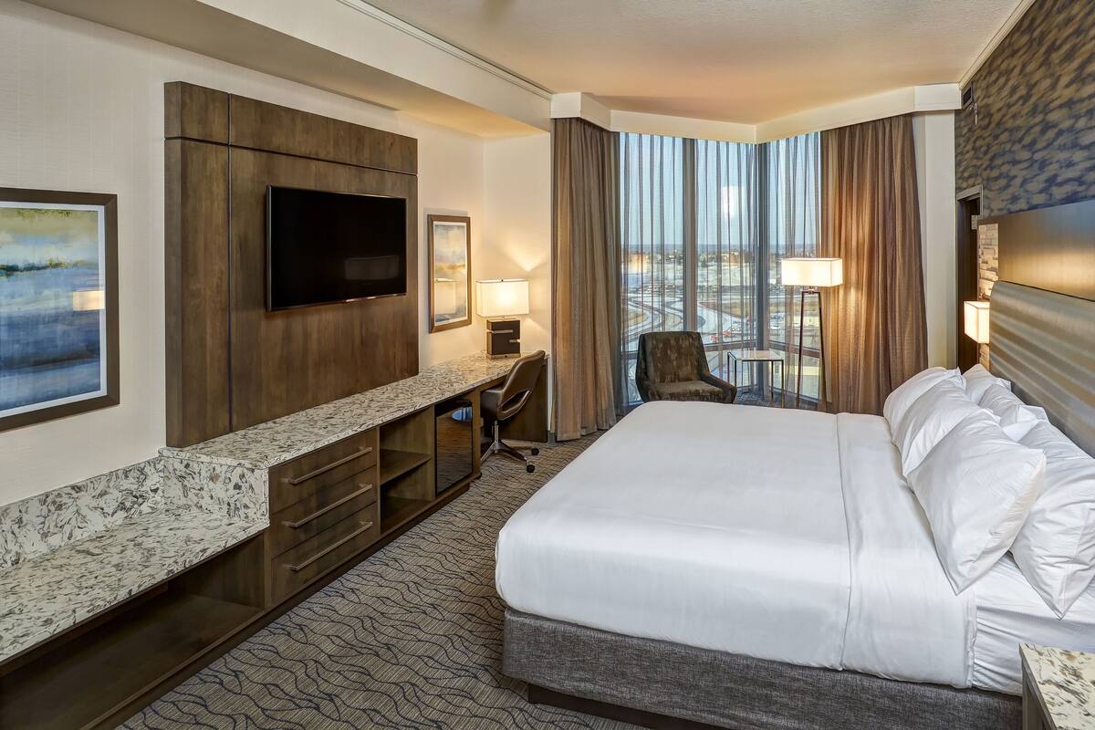 Ein Hotelzimmer mit einem großen Bett, einem an der Wand montierten Flachbildfernseher, einem Schreibtisch mit Stuhl, einem kleinen Tisch und Blick auf die Stadt durch große Fenster.
