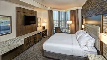 Quarto de hotel com uma cama grande, televisão de tela plana na parede, escrivaninha com cadeira, mesa pequena e vista da cidade por grandes janelas.