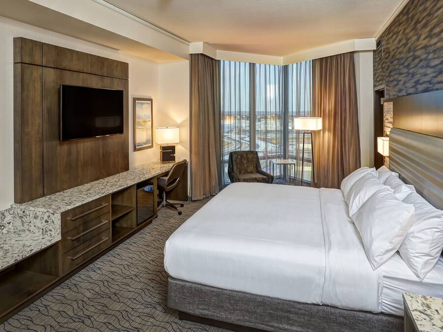 Quarto de hotel com uma cama grande, televisão de tela plana na parede, escrivaninha com cadeira, mesa pequena e vista da cidade por grandes janelas.