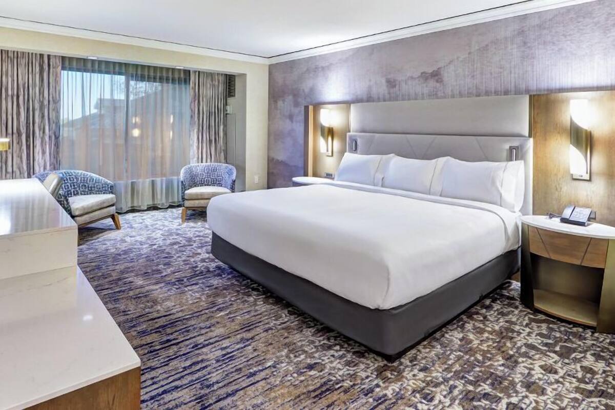 Ein modernes Hotelzimmer mit einem großen Bett, zwei Sesseln, einem Schreibtisch und einem Badezimmer mit Waschtisch.