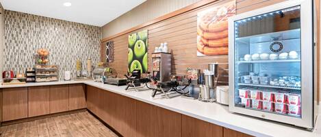 A breakfast bar with a refrigerator stocked with beverages, a coffee machine, and a display of pastries.