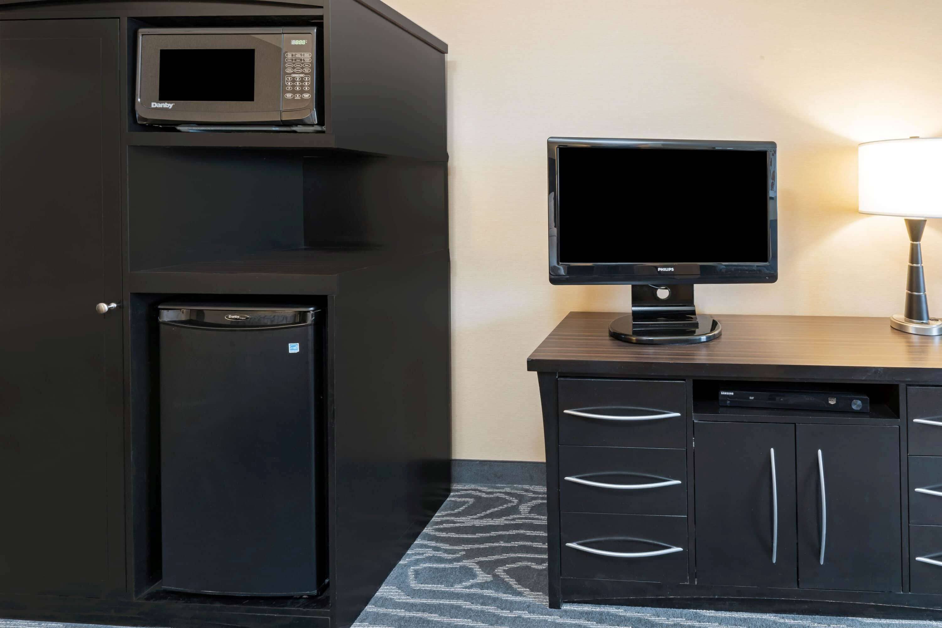 A compact kitchenette with a microwave, refrigerator, and a flat-screen TV on a wooden counter.