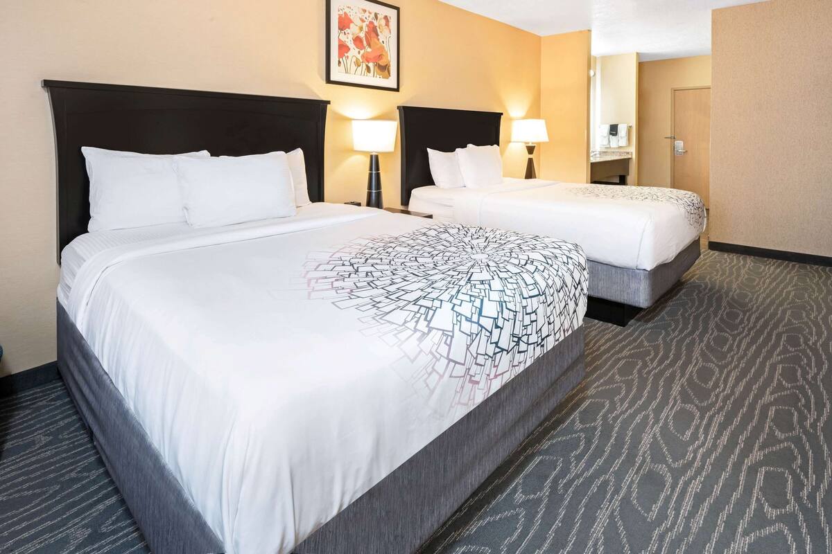 Ein Hotelzimmer mit zwei Betten, einem großen Bett mit einem gemusterten Überwurf und einem kleineren Bett mit schlichtem Bettbezug. Zwei Nachttischlampen, ein gerahmtes Bild an der Wand und eine Tür rechts sind ebenfalls zu sehen.
